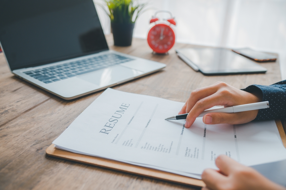Close-up of a person reviewing a printed resume with a pen at a wooden desk, featuring a laptop, red alarm clock, and tablet in the background.