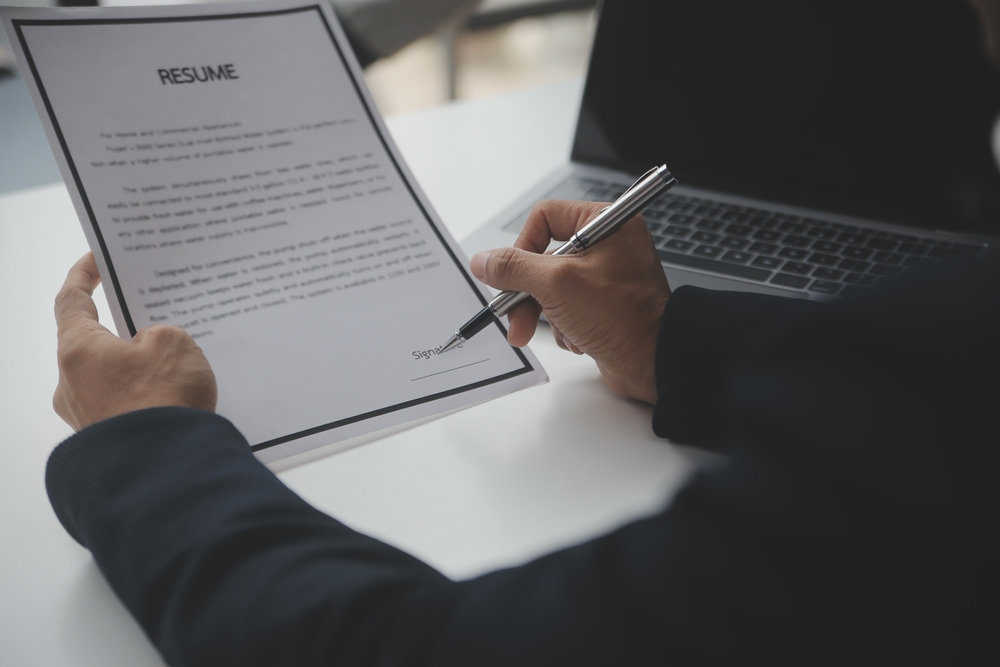 Person signing a printed resume at a desk beside an open laptop.