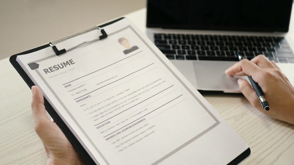 Person reviewing printed resume on clipboard beside open laptop.